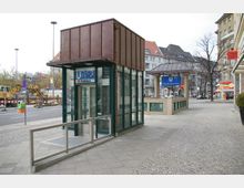 U-Bahnhof-Eingang in einer städtischen Umgebung mit modernen gläsernen Aufzugsschächten und Zugangstreppen, beschildert mit "U2 Ruhleben" und "U Theodor-Heuss-Platz". Im Hintergrund befinden sich mehrstöckige Wohnhäuser, Baustellenfahrzeuge und Geschäfte entlang einer Straße.