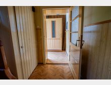 Schmaler Flur in einem älteren Wohnhaus mit Holzvertäfelung, Einbauschrank links und Parkett im Fischgrätmuster. Im Vordergrund steht eine halb geöffnete Tür mit strukturiertem Glas, die den Blick in einen angrenzenden Raum mit weiterer Innentür und Vorhang freigibt.
