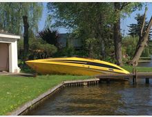 Gelbes Motorboot mit schwarzem Streifendesign, geparkt auf einer Rasenfläche neben einem kleinen Steg am Wasser; im Hintergrund sind Bäume, Büsche und ein weißes Gebäude mit einer offenen Garage zu sehen. Eine ruhige Umgebung mit einem Fluss im Vordergrund.