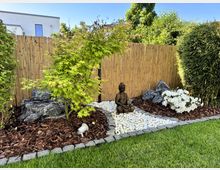 Ein kleiner Zen-Garten mit einem Boden aus weißen Kieselsteinen, dunkelbrauner Rindenmulch und grauen Steinen, umgeben von grünen Pflanzen. Eine Buddha-Statue sitzt in der Mitte, vor einem Sichtschutz aus Bambus; im Hintergrund ist ein modernes Gebäude erkennbar.