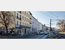 Straßenszene in einer Stadt mit mehrstöckigen, pastellfarbenen Wohnhäusern und Geschäften im Erdgeschoss. Rechts verlaufen Straßenbahnschienen und Oberleitungen, während links Autos und ein Radweg zu sehen sind, flankiert von einem Baum mit herbstlichem Laub.