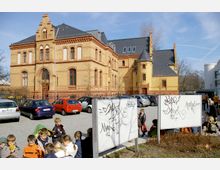Backsteinbau im Stil des Historismus mit verzierten Fensterbögen und Dächern, umgeben von einem gepflasterten Parkplatz mit mehreren Autos. Im Vordergrund stehen Kindergruppen sowie beschmierte Infotafeln.