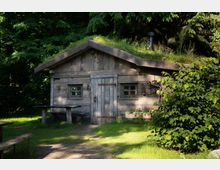 Blockhütte aus Holz mit Grasdach, umgeben von Bäumen und Büschen. Vor der Hütte befinden sich einfache Holzbänke und ein Tisch auf einer Wiese.