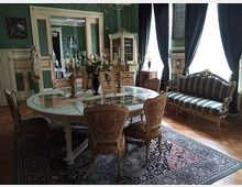 Eleganter Raum im klassischen Stil mit grün tapezierten Wänden, einem runden Esstisch mit reich verzierten Stühlen, einem dekorativen Teppich und einem gepolsterten Sofa mit Streifenmuster. Große Fenster mit dunklen Vorhängen lassen diffuses Licht einfallen, während Möbel und Dekorelemente wie Kerzenhalter und Blumenarrangements das Ambiente vervollständigen.