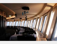 Restaurant mit Panoramafenstern in einem runden, modernen Gebäude. Der Raum ist mit gedeckten Tischen und Polsterstühlen eingerichtet, die Fenster lassen viel Tageslicht herein, während die Decke mit dezenten Dekorationen behängt ist.