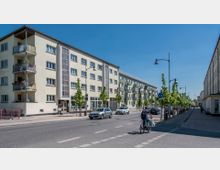 Straßenszene in einer Wohngegend mit mehrstöckigen, hell verputzten Wohngebäuden mit Balkonen. Entlang der Straße sind parkende Autos, vereinzelte Passanten, ein Radfahrer und eine Baumreihe auf dem Gehweg sichtbar.