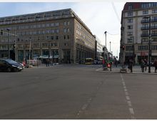 Grosser Straßenkreuzung in einer Stadt mit mehrstöckigen Gebäuden im Hintergrund, darunter ein Hotel und Geschäfte. Fußgänger, Autos und ein gelber Bus sind sichtbar, außerdem Ampeln und Straßenbaumaßnahmen an den Gehwegen.