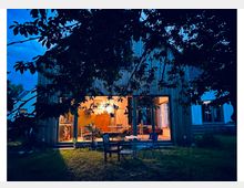 Holzhaus mit großem, bodentiefem Fenster zur Terrasse, durch das warm beleuchtete Innenräume sichtbar sind. Im Vordergrund liegen Rasenfläche und Gartenmöbel, eingerahmt von dunklen Baumzweigen in der Abenddämmerung.