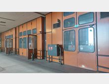 Industriehalle mit orangefarbenen Metallwänden und großen Fenstereinsätzen, teilweise mit Türen versehen. An der Wand befinden sich Sicherheitskästen mit Feuerlöschsymbolen, und der Boden ist aus Beton.