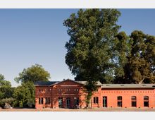 Ein einstöckiges Backsteingebäude mit großen Rundbogenfenstern, beschriftet mit „Seeterrassen“, umgeben von hohen Bäumen und einem gepflasterten Vorplatz unter klarem Himmel.