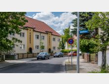 Wohnstraße mit zweistöckigen Mehrfamilienhäusern und roten Satteldächern, umgeben von Bäumen und Sträuchern. Im Vordergrund eine asphaltierte Straße mit parkenden Autos, sowie ein Straßenschild mit der Aufschrift „Bahnhofsplatz“.