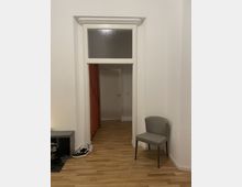 Innenraum mit weiß gestrichenen Wänden, einer hohen Tür mit Glaseinsatz im oberen Bereich sowie orangefarbenem Vorhang hinter der Tür. Der Boden besteht aus Holz, rechts steht ein grauer Stuhl, und links ist ein kleiner Schuhschrank mit Schuhen sichtbar.