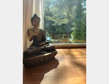 Bronzefarbene Buddha-Statue auf einem Holzboden direkt vor einer großen Glasfensterfront mit Vorhang. Durch das Fenster sind ein sonniger Garten mit Bäumen und ein Gewässer im Hintergrund zu sehen.