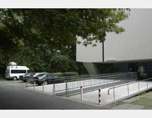 Modernes Gebäude mit überhängendem Obergeschoss, gestützt von großen V-förmigen Säulen. Im Vordergrund ein gepflasterter Gehweg, eine Rampe mit Geländer, sowie geparkte Autos vor einer Baumreihe.