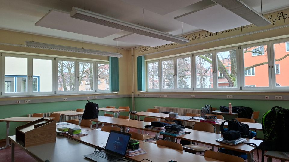 Klassenzimmer mit mehreren Reihen aus Holztischen und -stühlen sowie einer Lehrerpult-Ecke im Vordergrund. Entlang der Wand befinden sich breite Fensterfronten mit Blick auf Bäume und Wohngebäude; an der Decke hängen lange Leuchtstofflampen und Akustikplatten.
