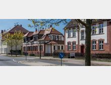 Straßenszene mit historischen Gebäuden, darunter ein Fachwerkhaus mit roten Dachziegeln und ein größeres Gebäude mit weißer Fassade und roten Klinkerelementen; im Vordergrund ein Baum und ein Verkehrszeichen auf einer Verkehrsinsel.