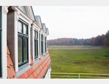 Fachwerkhaus mit schrägem Ziegeldach und weißen Fensterrahmen, angrenzend an eine weite Wiesenlandschaft mit eingezäunten Bereichen und herbstlichem Wald im Hintergrund.