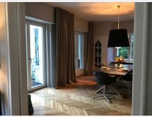 Esszimmer mit hellem Parkettboden, einem großen Holztisch und modernen schwarzen Stühlen. Große Fenstertüren mit beigefarbenen Vorhängen führen nach draußen, und ein großer schwarzer Lampenschirm hängt über dem Tisch.