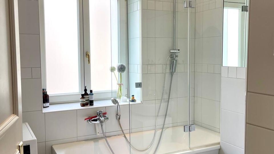 Helles, weiß gefliestes Badezimmer mit Badewanne und integrierter Duschwand aus Glas sowie Regendusche. Neben einem großen, hohen Fenster mit Rundbogen befinden sich eine Fensterbank mit Pflegeprodukten und rechts ein wandhängendes WC.
