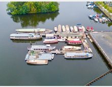 Luftaufnahme eines kleinen Hafens mit mehreren Anlegestellen, an denen verschiedene Boote und Yachten festgemacht sind. Im Hintergrund befindet sich eine bewaldete Insel, und das Wasser spiegelt die Umgebung.