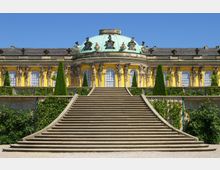 Gelbes barockes Schlossgebäude mit großen Fenstern und verzierten Figuren, gekrönt von einer grünen Kuppel. Im Vordergrund führen breite Steintreppen durch einen Gartenbereich mit geometrisch geschnittenen Pflanzen und Klettergewächsen.