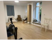 Heller Innenraum mit hellem Holzboden und weißen Wänden, eingerichtet mit einem Ruderergometer, einer flachen Trainingsbank und Sitzkissen. An der rechten Seite befindet sich ein großes Fenster mit Fensterbank-Sitzplatz sowie ein hoher Holzschrank und ein Stuhl.