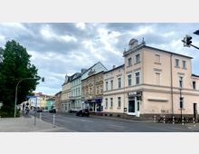 Straßenszene mit historischen Wohn- und Geschäftshäusern, darunter ein beigefarbenes Gebäude mit Pizzeria im Erdgeschoss. Links ein großer Baum und ein Straßenschild „An Kaiserhof“; im Hintergrund weitere bunte Gebäude unter bewölktem Himmel.