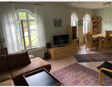 Wohnzimmer mit großen Fenstern, die mit weißen Vorhängen dekoriert sind, und einer geöffneten Flügeltür mit Blick auf einen grünen Garten. Im Raum befinden sich ein braunes Sofa, ein Fernseher auf einem Holzschrank, ein Esstisch mit Rattanstühlen, sowie gemusterte Teppiche auf einem Fliesenboden.