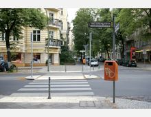 Straßenszene einer Wohngegend mit einem Zebrastreifen im Vordergrund, einem mehrstöckigen gelb beigen Gebäude mit Balkonen und Geschäften im Erdgeschoss links, sowie Bäumen, parkenden Autos und Straßenschildern im Hintergrund. Ein auffälliger orangefarbener Mülleimer steht auf der rechten Seite.