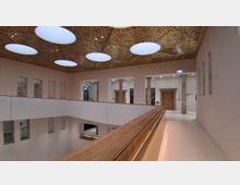 Innenansicht eines modernen Atriums mit umlaufender Galerie und hellem Geländer mit Holzhandlauf. Die Decke ist mit perforierten Paneelen und großen ovalen Oberlichtern gestaltet; entlang der Wände stehen Säulen und mehrere Türöffnungen.