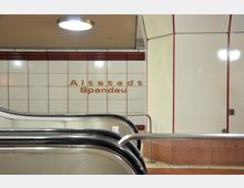 U-Bahn-Station mit weiß gefliesten Wänden, roten Zierlinien und der Aufschrift „Altstadt Spandau“. Im Vordergrund Rolltreppen und ein Metallgeländer.