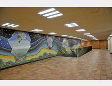 Unterirdischer Durchgang mit einer großen Wandmosaik-Kunst, die Weltraummotive wie Erde, Planeten, Sterne und ein Satellit zeigt. Die Wände und der Boden sind mit Holz- und Fliesenmaterialien gestaltet, und eine Treppe mit schwarzem Geländer führt nach unten.