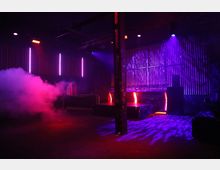 Ein dunkler Clubraum mit lila und roten Lichteffekten, einer Bühne mit metallischen Gitterelementen und vertikalen Lichtleisten an den Wänden. Im Vordergrund ist eine Rauchwolke zu sehen, und die Szene wirkt durch die Beleuchtung und Schatten atmosphärisch.