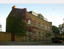 Backsteingebäude aus der Gründerzeit mit verzierten Fassaden und mehreren Reihen weißer Fenster, dessen Seitenwand teilweise mit grünem und rotem Efeu bewachsen ist. Im Vordergrund eine gepflasterte Straße, eine niedrige Backsteinmauer mit Holztor und ein geparktes Auto.