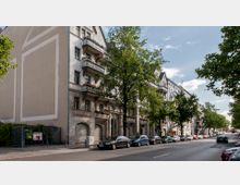 Straßenansicht einer Wohnstraße mit historischen Mehrfamilienhäusern, Balkonen und dekorativen Fassaden. Am Straßenrand stehen geparkte Autos, während Bäume den Gehweg säumen.