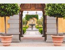 Ein gepflegter Innenhof mit einem zentralen Springbrunnen, eingerahmt von symmetrisch geschnittenen Hecken und Bäumen in Terrakotta-Töpfen, sichtbar durch ein offenes Holztor in einer gelb verputzten Fassade mit grauen Steinelementen. Im Hintergrund ist eine mit grünem Efeu bewachsene Wand mit einem architektonischen Bogen zu sehen.