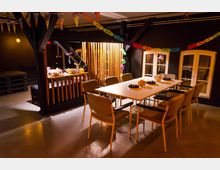 Ein festlich dekorierter Raum mit einem langen weißen Tisch, umgeben von Stühlen, und Girlanden, die an der Decke hängen. Im Hintergrund befindet sich eine Theke mit Snacks und Getränken sowie zwei große Fenster mit weißen Rahmen.