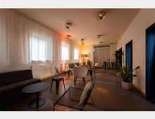 Ein moderner Lounge-Bereich mit minimalistischer Einrichtung, darunter Sofas, Stühle und kleine Tische. Der Raum ist von großen Fenstern mit weißen Vorhängen und warmem, indirektem Licht beleuchtet, mit dekorativen Pflanzen und einem schlichten, funktionalen Design.