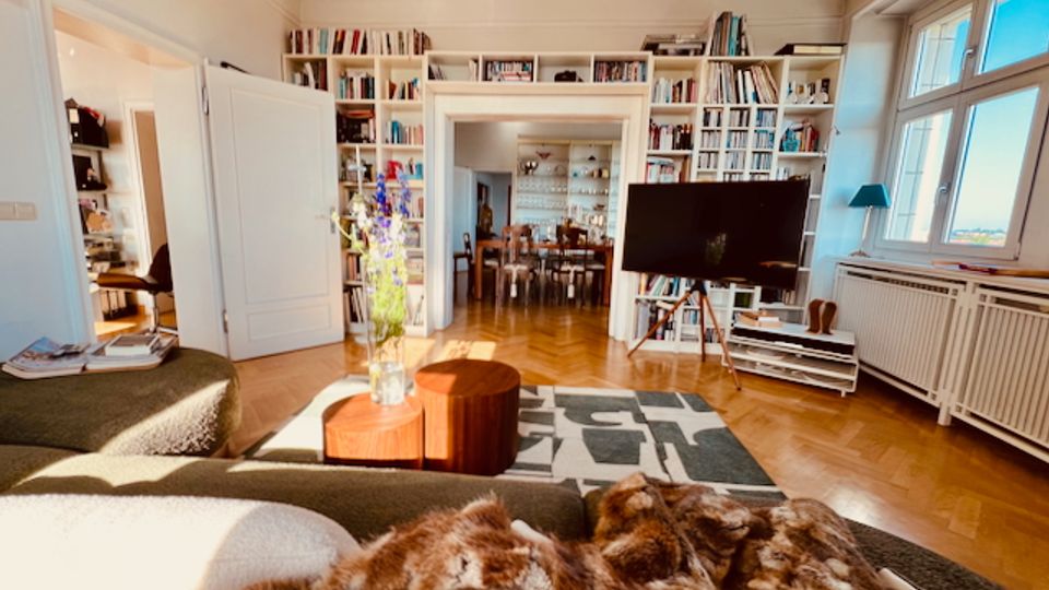 Helles Wohnzimmer mit Parkettboden, großem Fenster und deckenhohen Einbauregalen voller Bücher entlang der Wände. Im Raum stehen ein Sofa, niedrige Couchtische, ein Teppich sowie ein Fernseher; durch eine breite Türöffnung ist ein angrenzendes Esszimmer sichtbar.