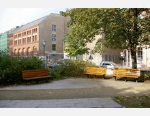 Kleiner Stadtpark mit gepflasterten Wegen, drei Holzbänken und umgeben von Sträuchern und Bäumen. Im Hintergrund ein historisches Backsteingebäude, moderne Häuser und parkende Autos.