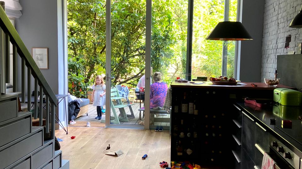 Offener Wohn‑ und Küchenbereich mit Holzfußboden, Treppe links und Küchenzeile rechts an einer weißen Ziegelwand. Große Schiebetür führt auf eine kleine Terrasse mit Gartenblick; im Raum liegen Spielzeug und Bausteine auf dem Boden.