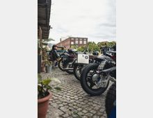 Mehrere Motorräder sind auf einem gepflasterten Hof geparkt, daneben stehen Pflanzen in Blumentöpfen. Im Hintergrund ist ein rotes Backsteingebäude sowie Bäume zu sehen, und eine Person sitzt auf einem Stuhl am Rand des Hofes.