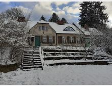 Ein eingeschossiges Wohnhaus mit schneebedecktem Satteldach, alten Fensterläden und grün gestrichenen Türen. Vor dem Gebäude führt eine Treppe durch einen winterlichen Garten mit verschneiten Sträuchern und Bäumen.