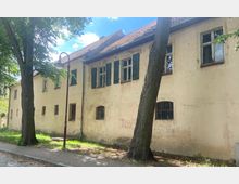 Zweistöckiges, verputztes Gebäude mit verblasster Fassade und klassischen Fenstern, teils mit grünen Fensterläden, darunter vergitterte Fenster; umgeben von Bäumen und mit einem gepflasterten Gehweg davor.