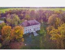 Luftaufnahme eines Herrenhauses mit hellbeiger Fassade und roten Dachziegeln, umgeben von herbstlich verfärbten Bäumen in einem weitläufigen Park. Im Hintergrund sind grüne Felder und eine flache Landschaft zu sehen.
