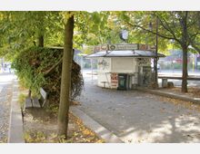 Ein kleiner Imbissstand mit der Aufschrift "Asia Snack" steht auf einem gepflasterten Platz, umgeben von Bäumen und fallendem Laub. Neben dem Stand befindet sich eine Sitzbank sowie ein Abfallcontainer, und Graffiti bedecken Teile des Gebäudes.