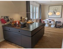 Offene Wohnküche mit großer dunkler Kücheninsel und integrierter Kochfläche, daneben eine Wand mit aufgehängten Küchenutensilien. Im Hintergrund ein Essbereich mit langem Tisch, gemischten Stühlen, zwei runden Pendelleuchten, großem Wandbild sowie hohen Fenstern mit Vorhängen über Fischgrätparkett.