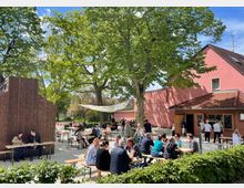 Ein Biergarten mit einer lockeren, sommerlichen Atmosphäre. Mehrere Menschen sitzen an Holzbänken unter großen Bäumen und vor einem rosa Gebäude mit Kioskfenstern und Speisetafeln.