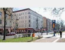 Straßenszene in einem urbanen Viertel mit mehrstöckigen Altbauten, Geschäften und Cafés im Erdgeschoss. Im Vordergrund befinden sich eine Parkbank, ein grüner Bereich mit Büschen und Bäumen sowie ein blaues Uhrenschild, während Fußgänger und Radfahrer auf der Straße und am Gehweg unterwegs sind.