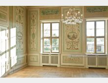 Eleganter historischer Innenraum mit pastellfarbenen Wandmalereien, die Landschaftsmotive und florale Verzierungen zeigen. Zwei große Fenster mit weißen Holzrahmen lassen Tageslicht herein, und ein Kristallleuchter hängt von der Decke über dem Parkettboden.